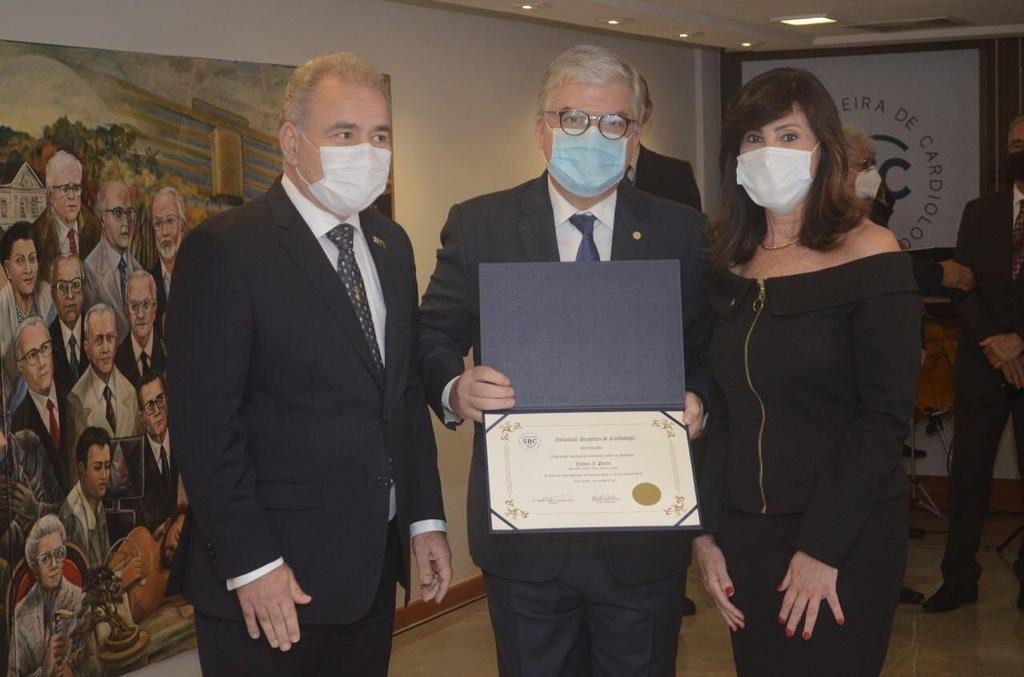 dois homens e uma mulher, todos de máscara, o homem que está no centro segura um diploma