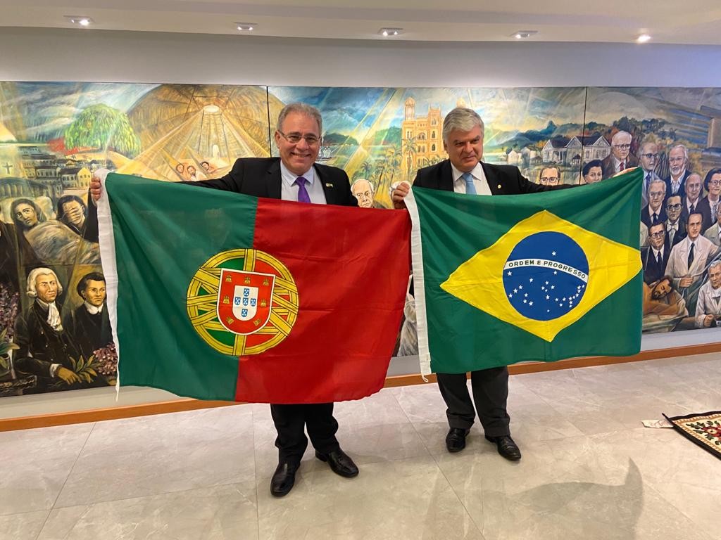 dois homens lado a lado, um segura a bandeira de Portugal, o outro a bandeira do Brasil