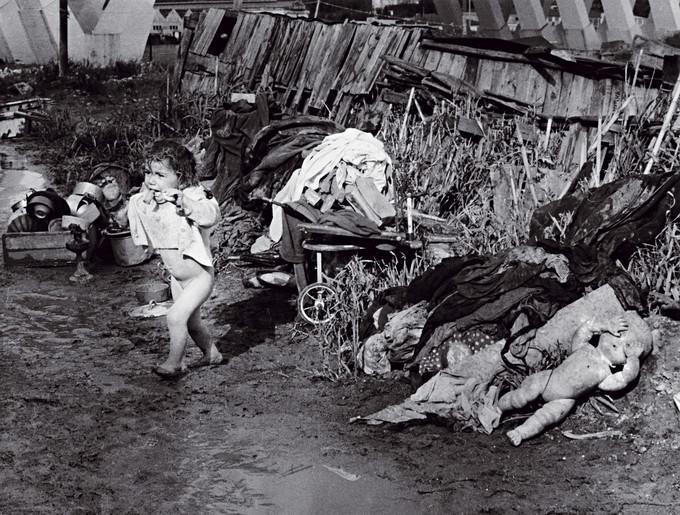 Criança despida e assustada com os pés na lama rodeada de detritos e lixo