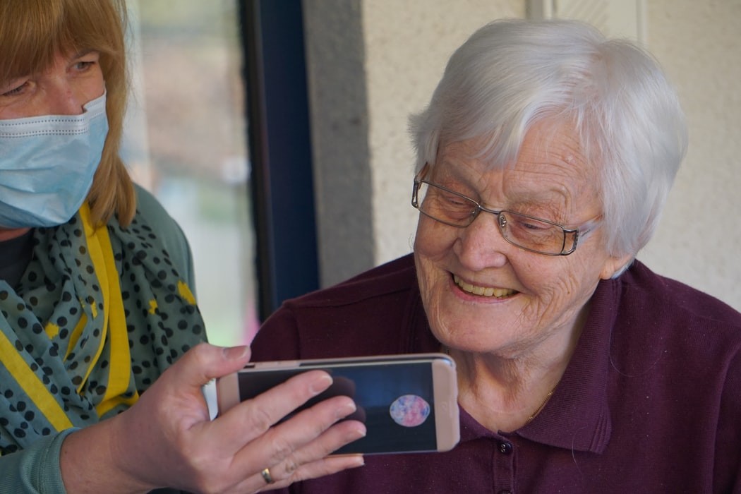 senhora de máscara segura telemóvel para que uma idosa possa comunicar por vídeo-chamada