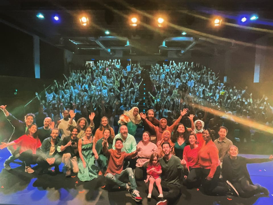 grupo de pessoas em cima do palco tiram selfie com a plateia em plano de fundo